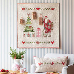 Decorative quilt with Christmas design hanging on a wall above a white armchair.