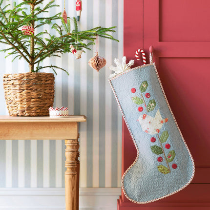 Decorative Christmas stocking hanging on a red door next to a small tree and table.